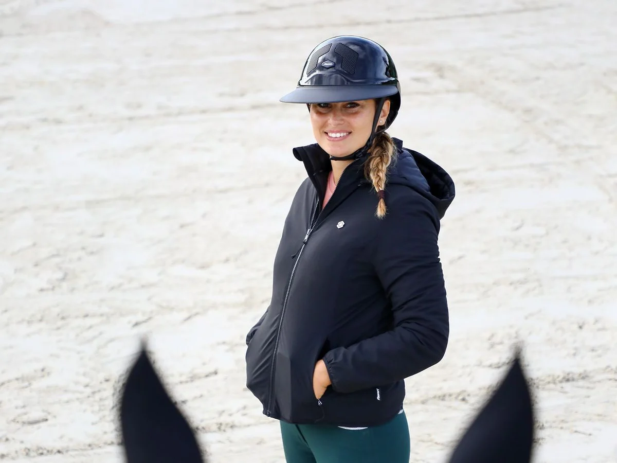 A rider wears a fitted jacket, riding trousers and helmet at an outdoor arena.