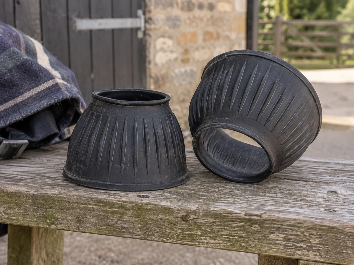 Pair of plain black bell-shaped rubber overreach boots with ribbed outer walls and circular openings on a low wooden bench.