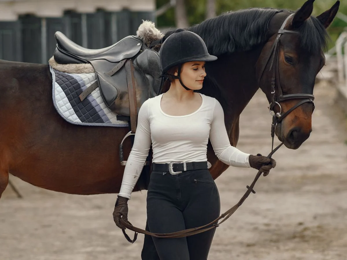 A rider in a light long-sleeved top, breeches, gloves and riding hat beside a horse.