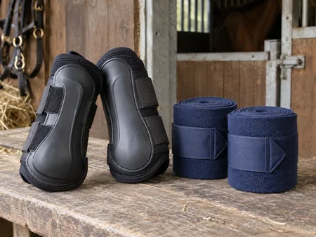 A pair of black horse brushing boots with shaped protective panels alongside two neatly rolled navy fleece exercise bandages on a stable bench.