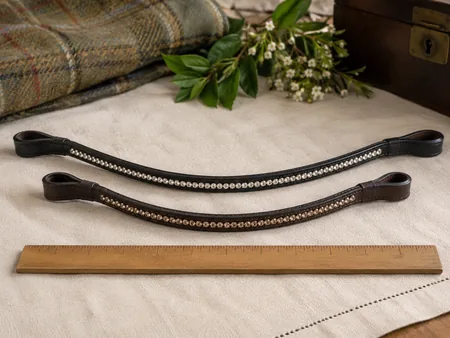 Two leather bridle browbands of different lengths arranged in shallow arcs on a cream cloth.