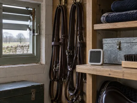 A small digital hygrometer with indistinct screen on a tack room shelf beside a slightly open vent and neatly stored leather reins.