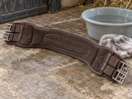 The underside of a detached brown horse girth turned upward beside a soft damp cloth and plain basin.