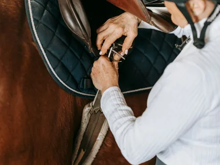 A rider checks the girth buckles beneath an English saddle flap.