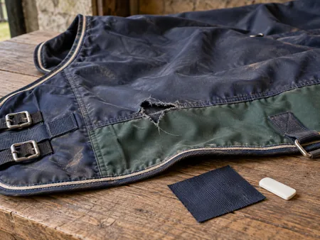 A detached horse rug with a small torn section laid flat beside a fabric repair patch and tailoring chalk.
