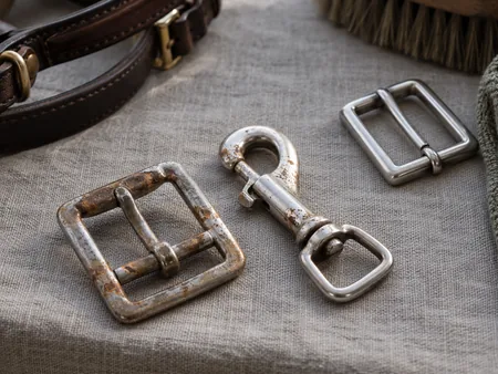 Close view of a removed tack buckle and clip with light rust spots beside a clean stainless buckle on grey linen.