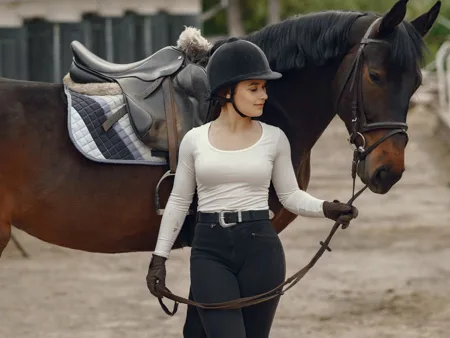 A rider in a light long-sleeved top, breeches, gloves and riding hat beside a horse.