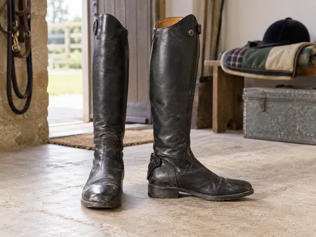 A pair of used black tall riding boots standing on a pale floor.